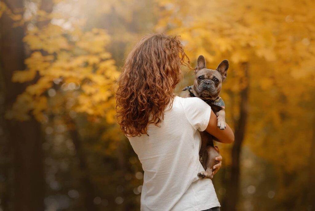 Photo of a woman standing outside against fall leaves holding her french bulldog, the bulldog is looking over her shoulder at the camera